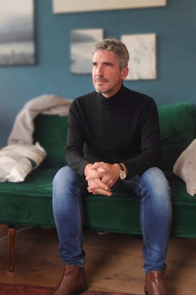 Homme assis sur un canapé vert, portant un pull noir et jeans, regard pensif.