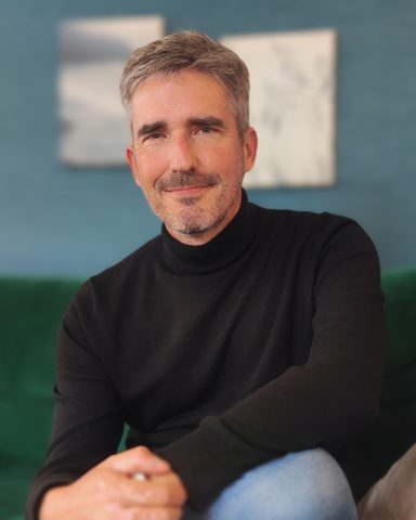 Homme avec un pull noir, assis sur un canapé vert, souriant à l'appareil photo.