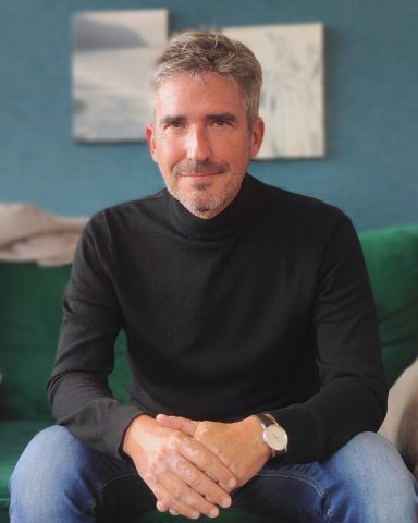 Homme souriant en pull noir, assis sur un canapé vert, avec un décor moderne en arrière-plan.